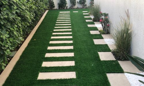 Narrow side garden design with artificial green grass and modern rectangular stepping stone pathway, bordered by green hedge and decorative plants - side yard landscaping Dubai