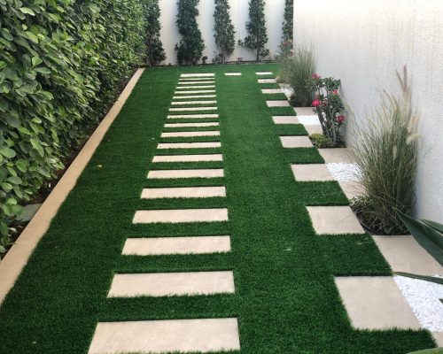 Narrow side garden design with artificial green grass and modern rectangular stepping stone pathway, bordered by green hedge and decorative plants - side yard landscaping Dubai
